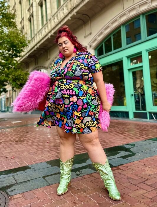 A person with red hair wears a colorful patterned dress, metallic green boots, and holds a pink furry accessory, standing on a brick sidewalk in front of a building with green trim.
