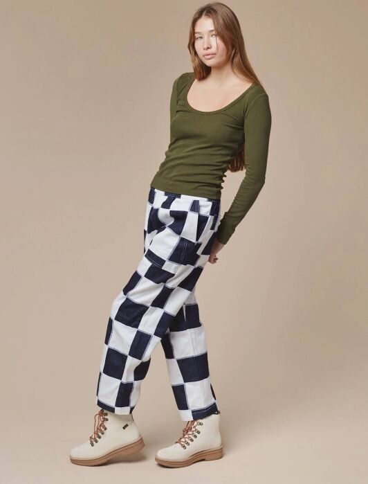 A person stands against a beige background, wearing an olive green long-sleeve top, black and white checkered pants, and beige boots.