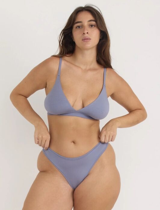 A woman stands against a plain background wearing a matching light purple bra and high-waisted underwear set, looking straight at the camera.