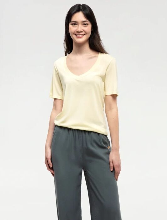 A woman stands smiling, wearing a light yellow short-sleeve shirt and dark gray pants against a plain light background.
