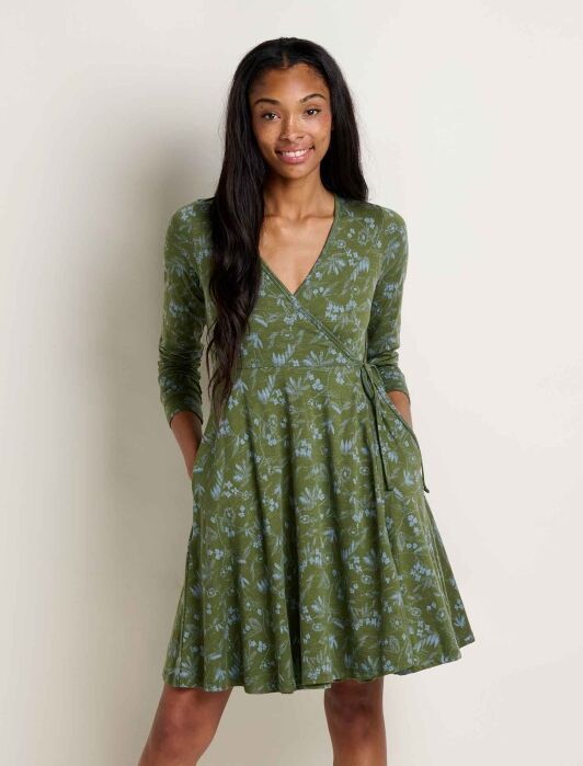 A woman stands against a plain background wearing a long-sleeve, green wrap dress with a subtle light blue floral pattern and pockets.