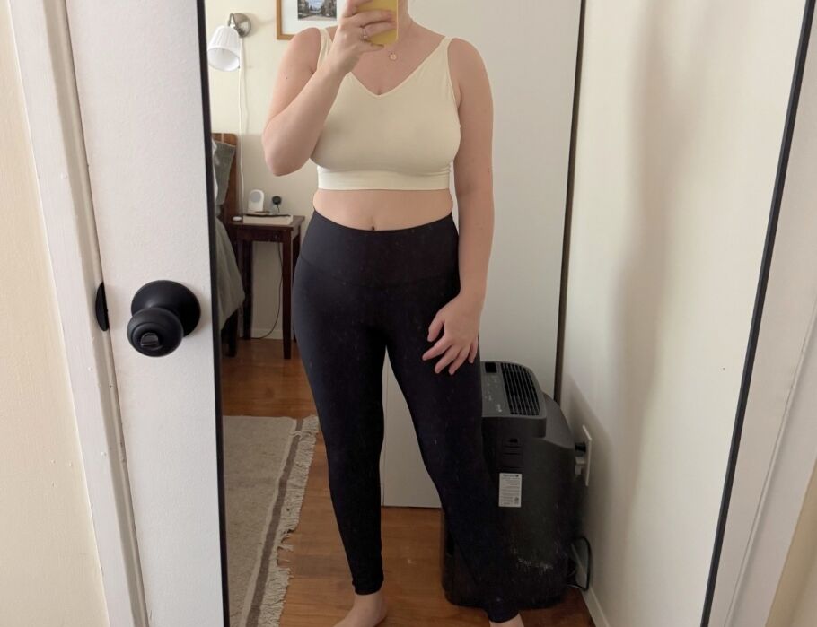 A person stands barefoot in front of a mirror wearing a white crop top and black leggings, with one hand on their hip. A room with a bed and small heater is visible in the background.