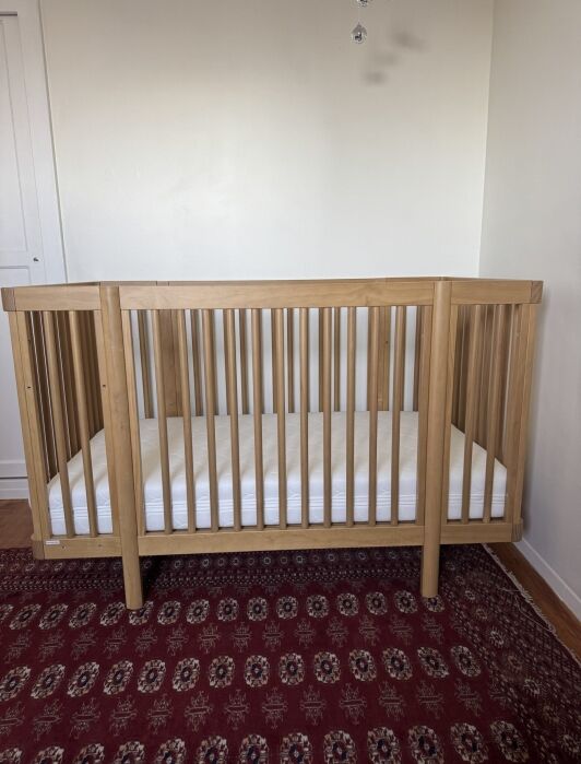 A wooden baby crib with vertical slats and a white mattress sits on a red patterned rug against a plain white wall.