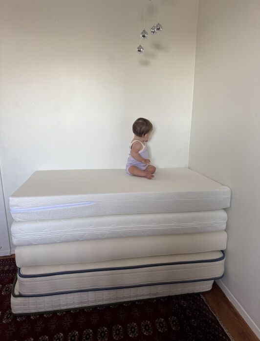 A small child sits on top of a stack of four mattresses placed in the corner of a room with white walls and a red carpet.
