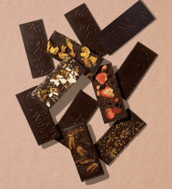 Assorted dark chocolate bars, some topped with nuts, dried fruit, cornflakes, or sprinkles, displayed on a pink textured surface.